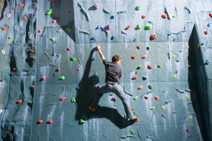 Picture of a climbing wall, with a man wearing jeans and a grey t-shirt climbing up it.