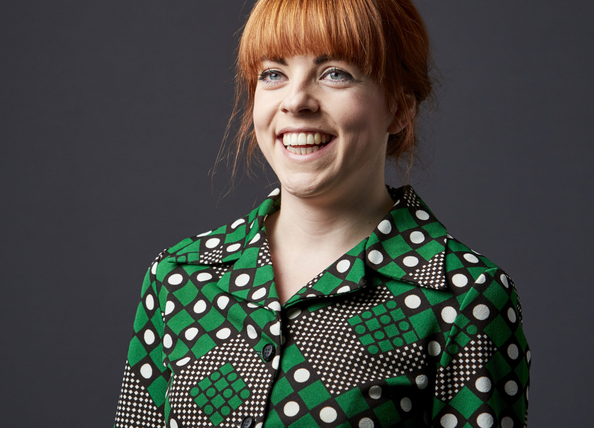 The picture shows a red headed woman, wearing a green, black and white shirt, smiling.