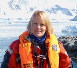 A photograph of Professor Dame Jane Francis. She is a white woman with blonde hair ab wearing a life jacket in front of a glacier.